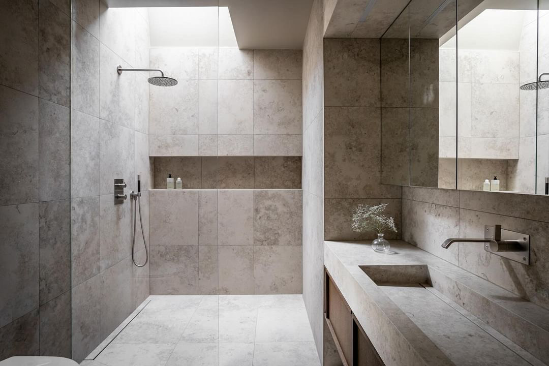 Elegant badrum med modern dusch och stilsäker inredning i naturinspirerade toner. Väggar och golv i ljus stenfinish, infällda hyllor för förvaring och integrerad spegel över ett minimalistiskt handfat.