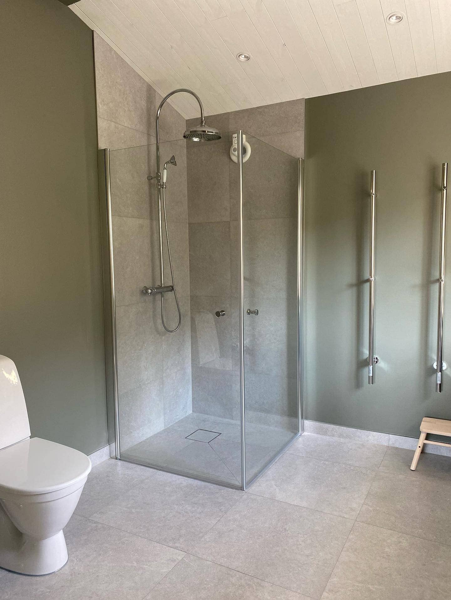 Bild på del av ett badrum med stora naturfärgade plattor på golvet och i duschhörnan. Gröna väggar, träpanel i taket.