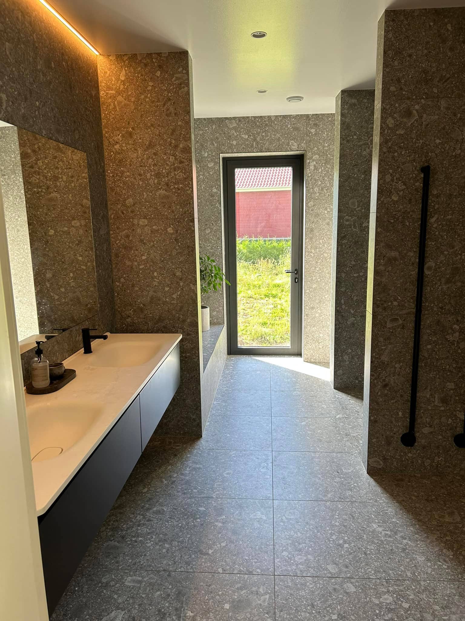 Badrum i sten med stora plattor som går i brunt. Vy där man ser handfat till vänster och en glasdörr som vetter ut rakt fram.