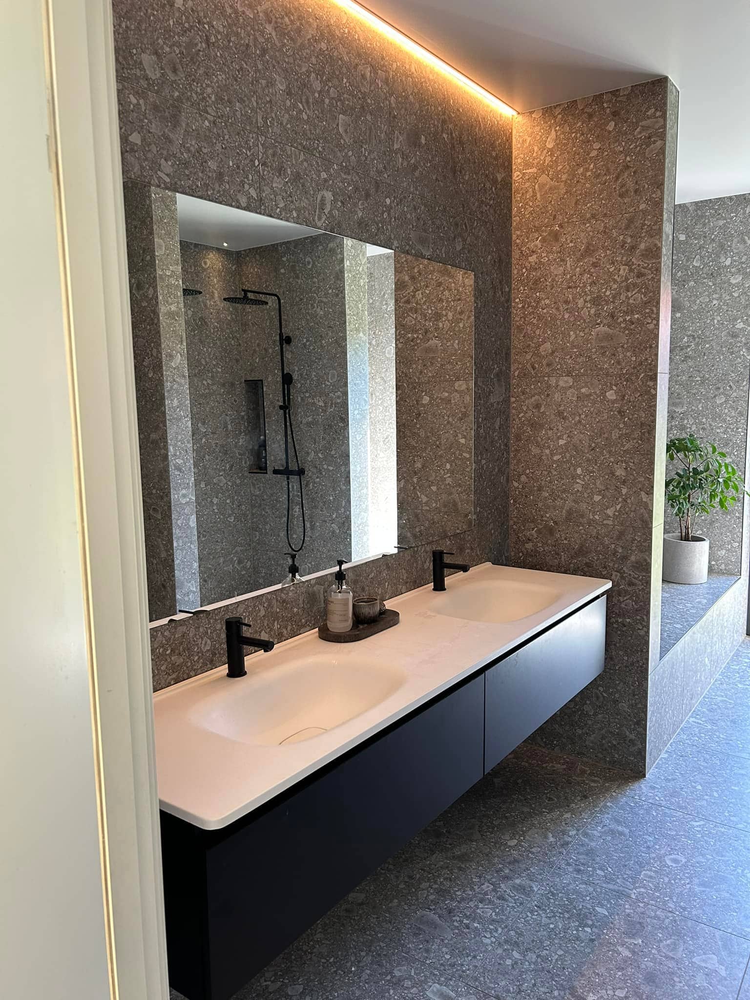 Modernt badrum med stora stenplattor i brun ton. På bilden syns en stor spegel som täcker väggen över det dubbla handfatet. Man skymtar en stennisch som kommer efter handfaten.