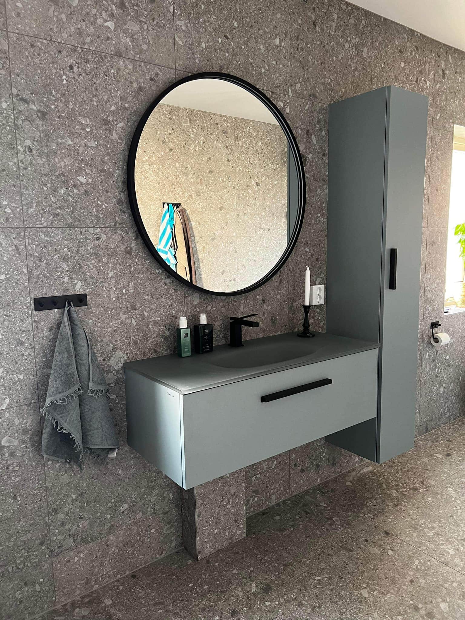 Modernt badrum med stora stenplattor i grå ton. I fokus på bilden är väggen med handfatet,rund spegel över och väggskåp till höger om handfatet.