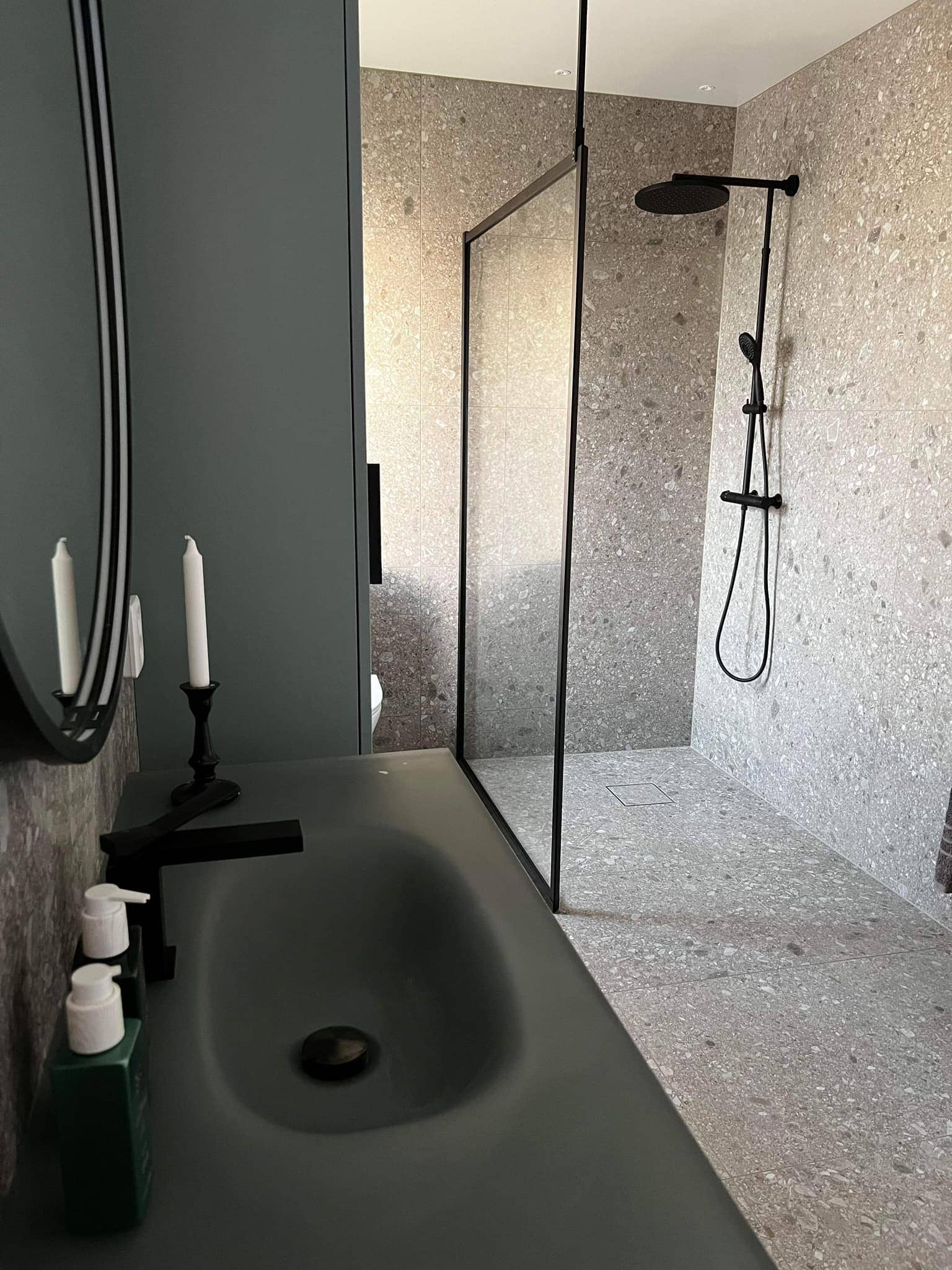 Duschutrymme i modernt badrum med svart dusch, duschvägg i glas och stora stenplattor i grå ton. till vänster i bild ser man även del av handfatet och ett smalt högskåp i grön färg.